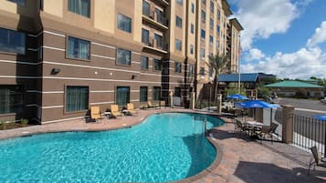 A hotel with a swimming pool, outdoor seating, and a clear sky.