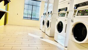 A laundry facility with multiple washing machines and dryers, a tiled floor, and a window with blinds.