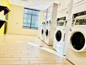 A laundry facility with multiple washing machines and dryers, a tiled floor, and a window with blinds.