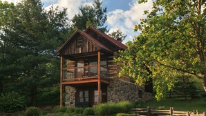 A two-story log cabin with a balcony, surrounded by a stone foundation and a wooden fence.