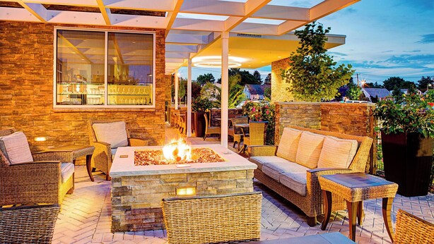 An outdoor patio area with wicker furniture, a stone fire pit, and a glass-enclosed bar. - Springhill Suites by Marriott Carle Place Garden City (Carle Place)