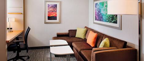 A hotel room with a brown sofa, two white tables, a desk with a chair, and two framed artworks on the wall.