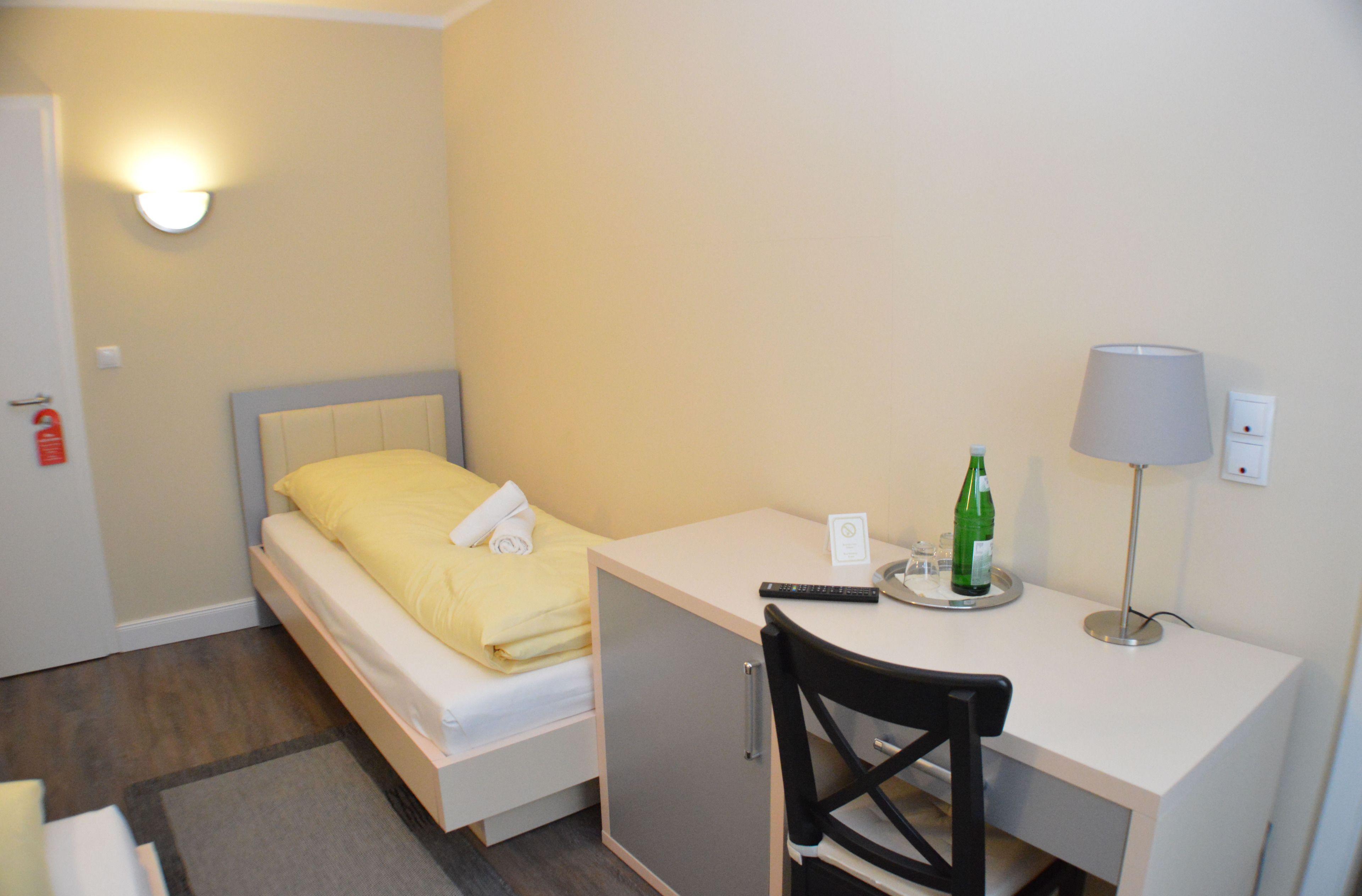 Une petite chambre avec un lit simple, un bureau, une chaise, une lampe et une bouteille posée sur le bureau.
