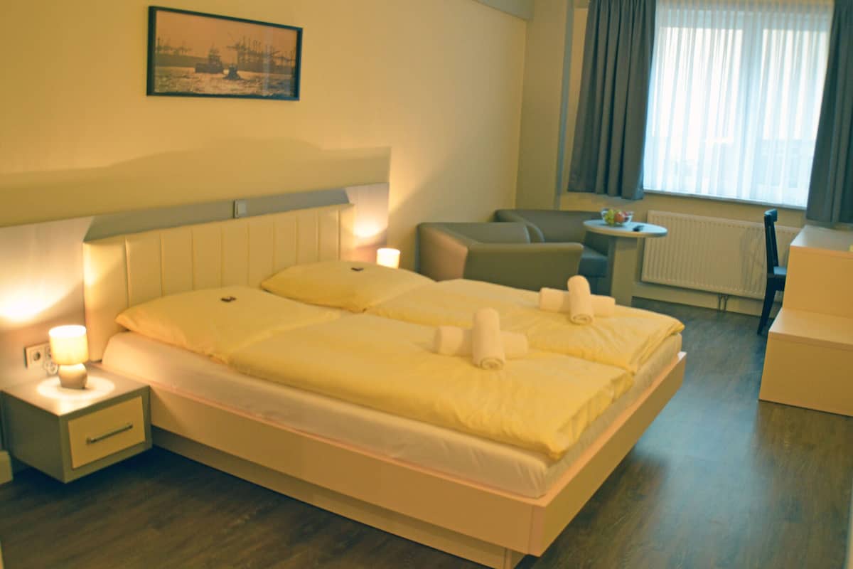 Ein Hotelzimmer mit einem großen Bett, zwei Nachttischen, einem kleinen Tisch, einem Sessel und einem Fenster mit Vorhängen.