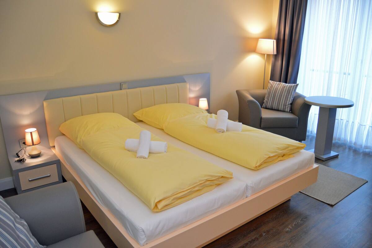Ein Hotelzimmer mit zwei Betten, jeweils mit gelber Bettwäsche und weißen Handtüchern, einem Nachttisch mit Lampe, einem kleinen Stuhl und einem runden Tisch.
