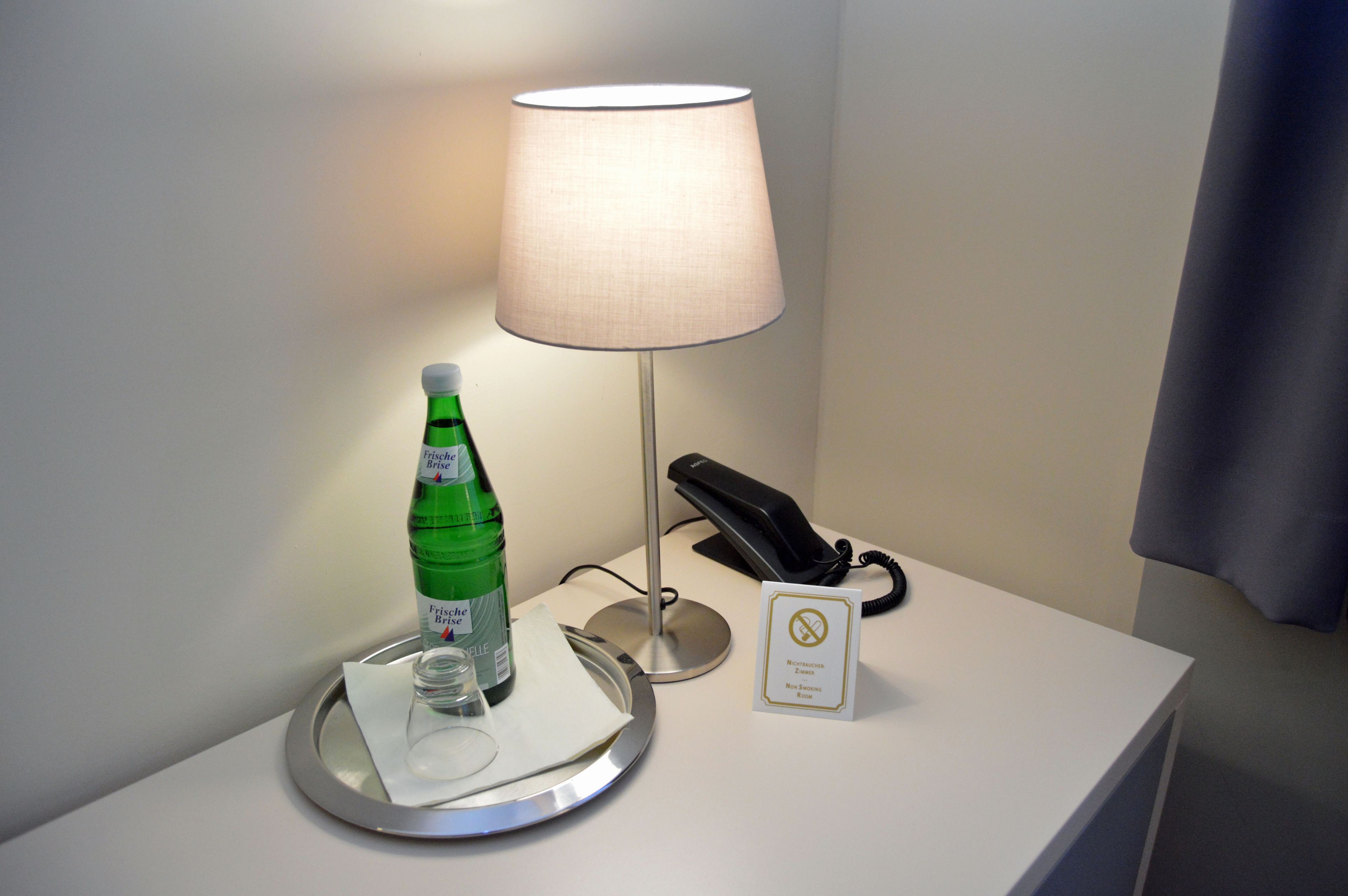 Een hotelkamer met een nachtkastje met een lamp, telefoon, een fles water en een glas.