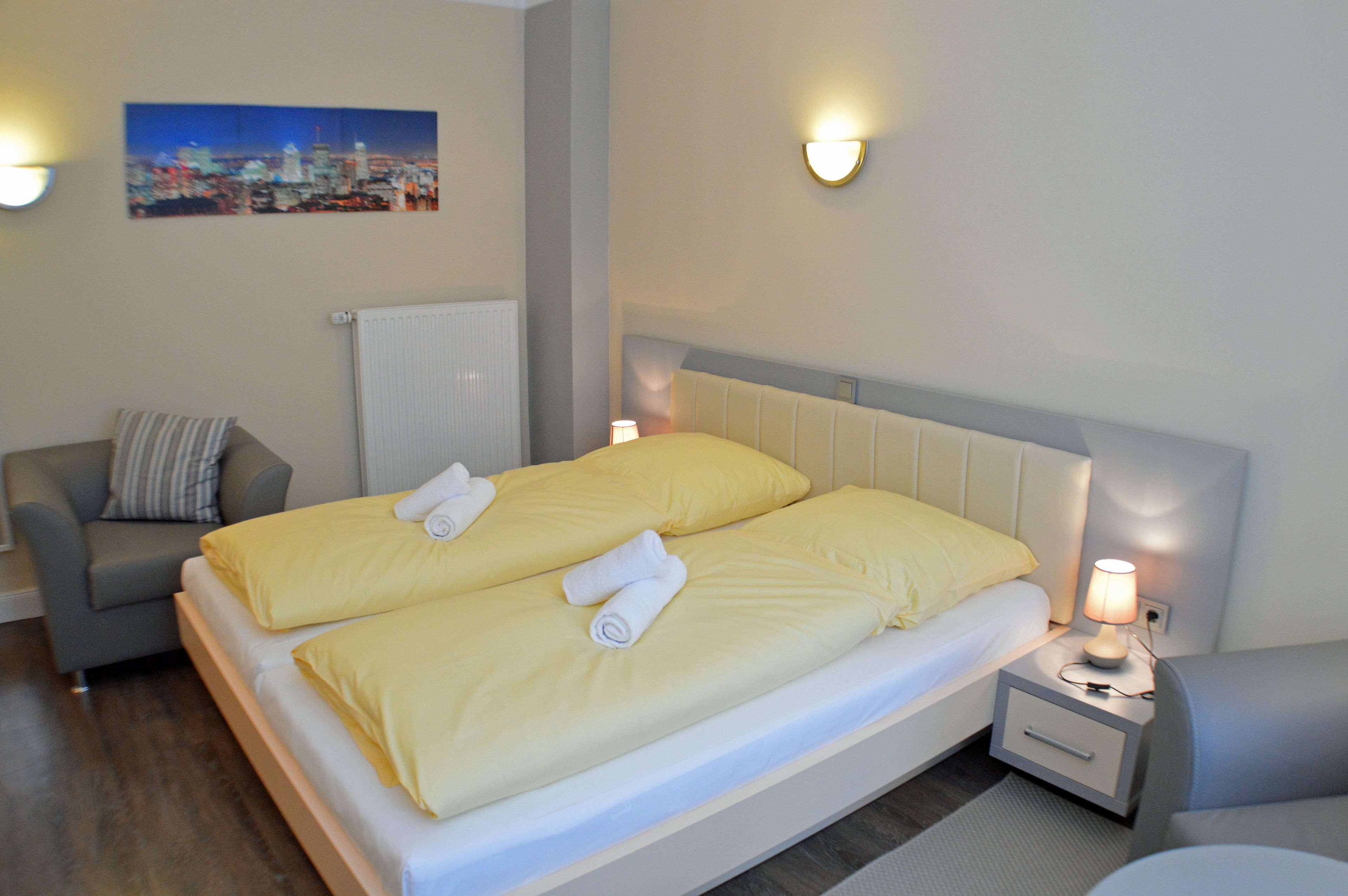 A hotel room with a double bed, a grey armchair, a bedside table with a lamp, and a cityscape painting on the wall.