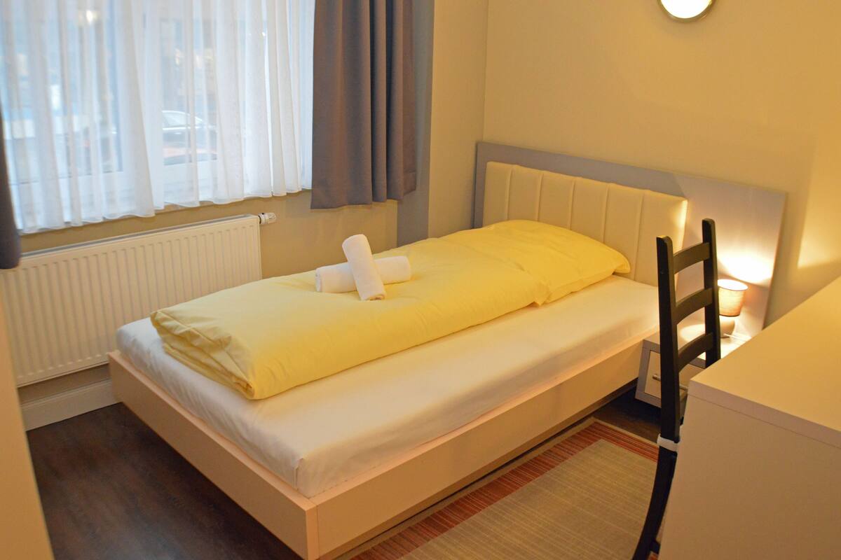 Ein Einzelbett mit weißem Kopfteil und gelber Decke. Darunter befindet sich unter dem Fenster eine weiße Heizung, und ein Lampenschirm steht auf dem Nachttisch.