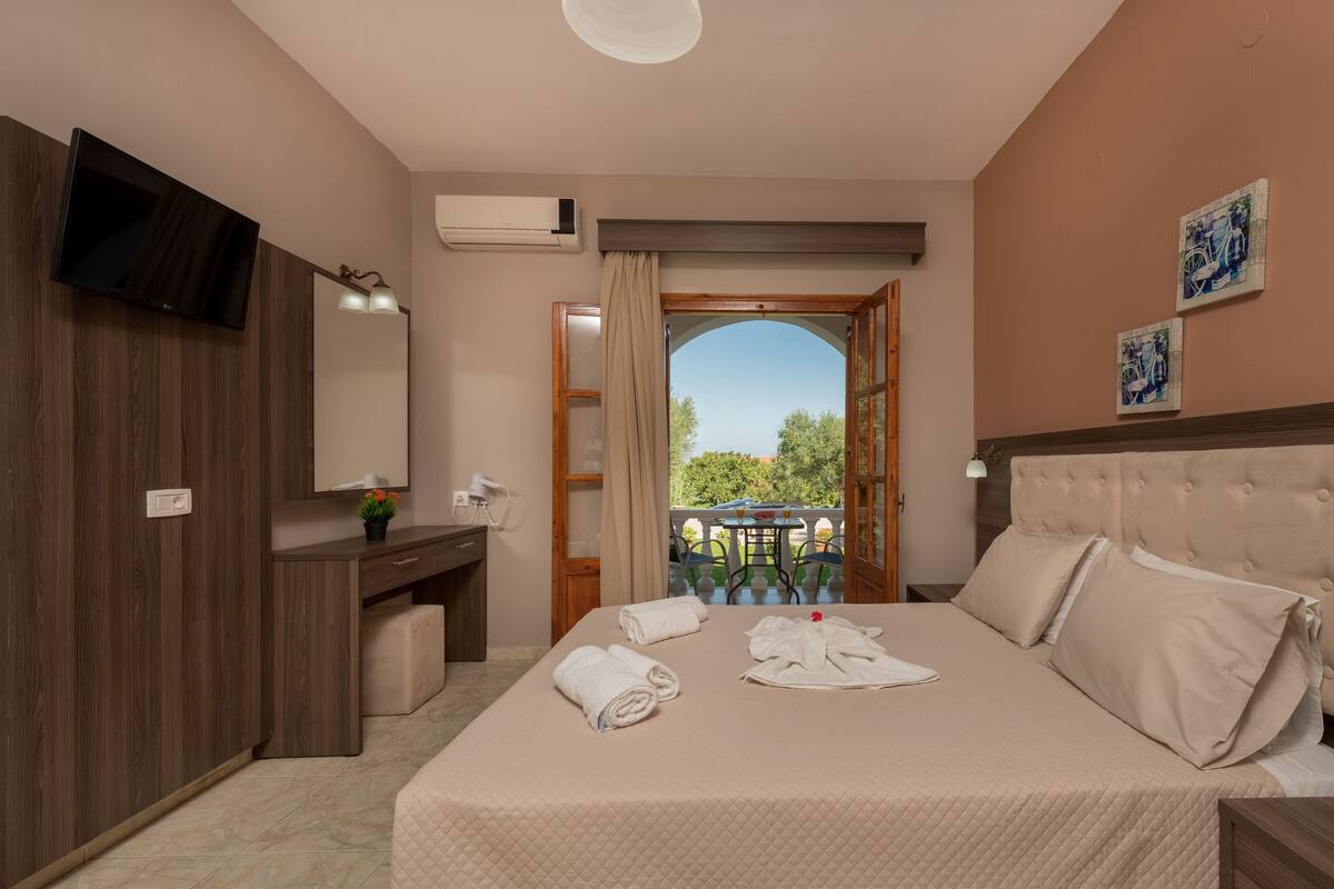 A hotel room with a bed, a desk, a mirror, an air conditioner, and a view of the outdoors.