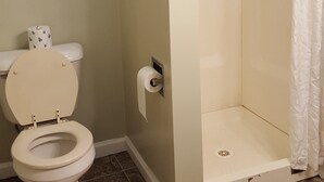 A bathroom with a toilet, a shower, and a toilet paper holder. - Colonial West Motel (Batavia)