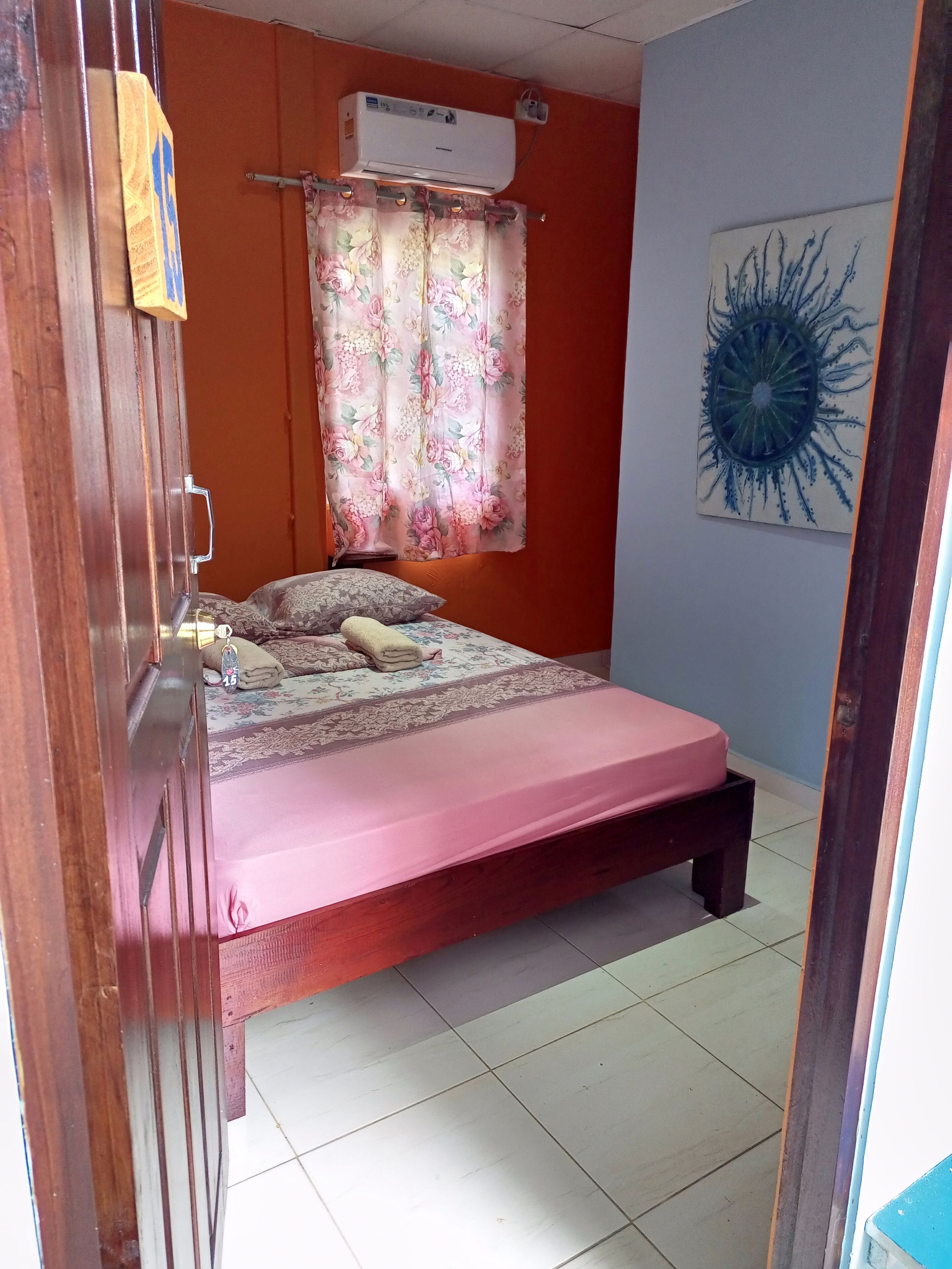 A bedroom with a bed, a window with floral curtains, an air conditioning unit, and a wall-mounted artwork.