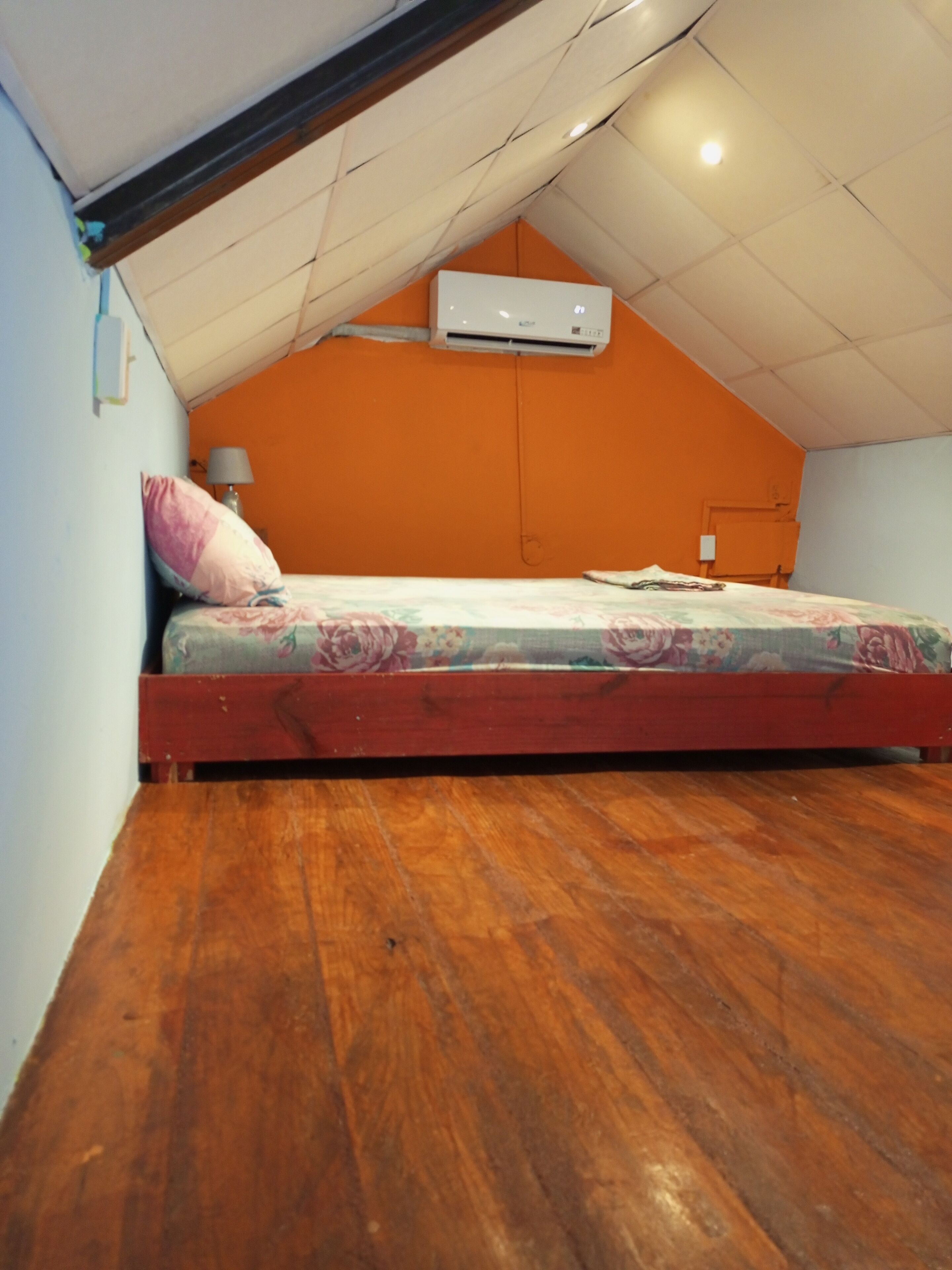 Un dormitorio con techo inclinado, una cama individual con ropa de cama floral, un equipo de aire acondicionado y piso de madera.