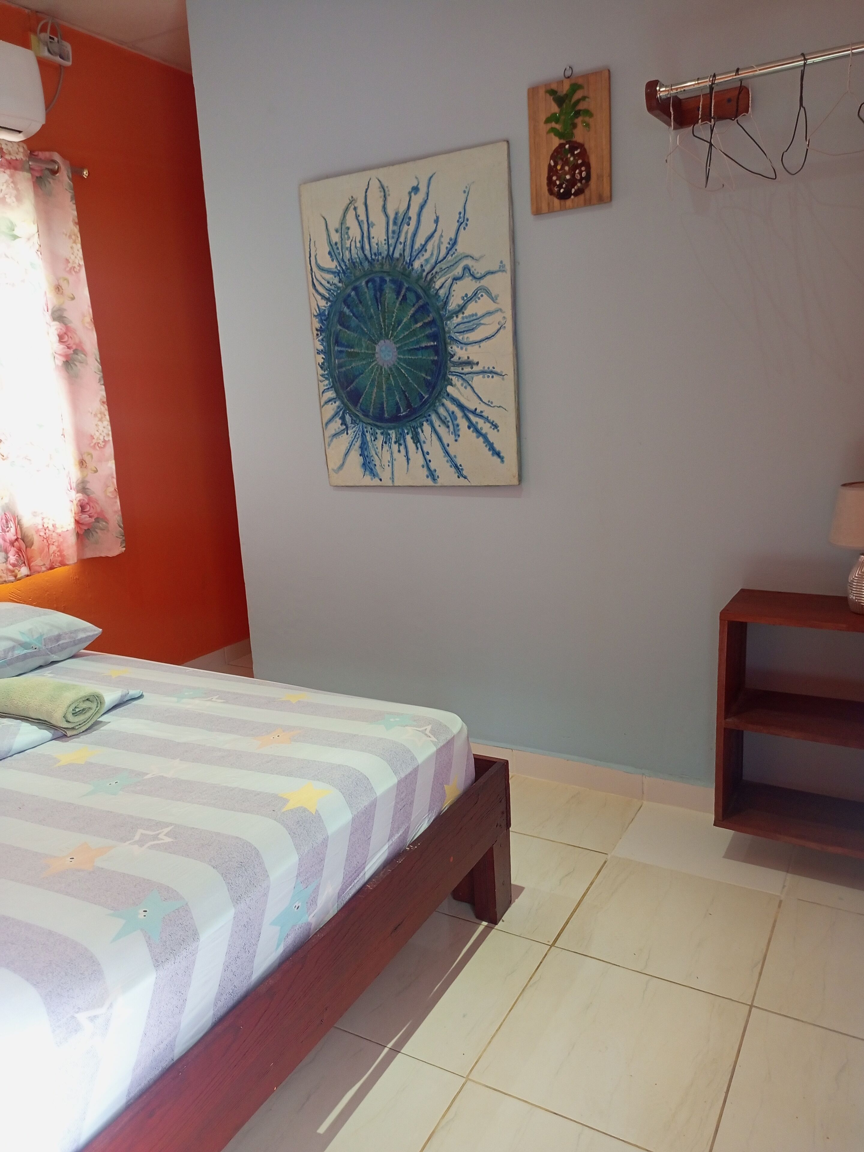 A bedroom with a bed, a wall-mounted air conditioner, a painting, a wooden shelf, and a window with curtains.