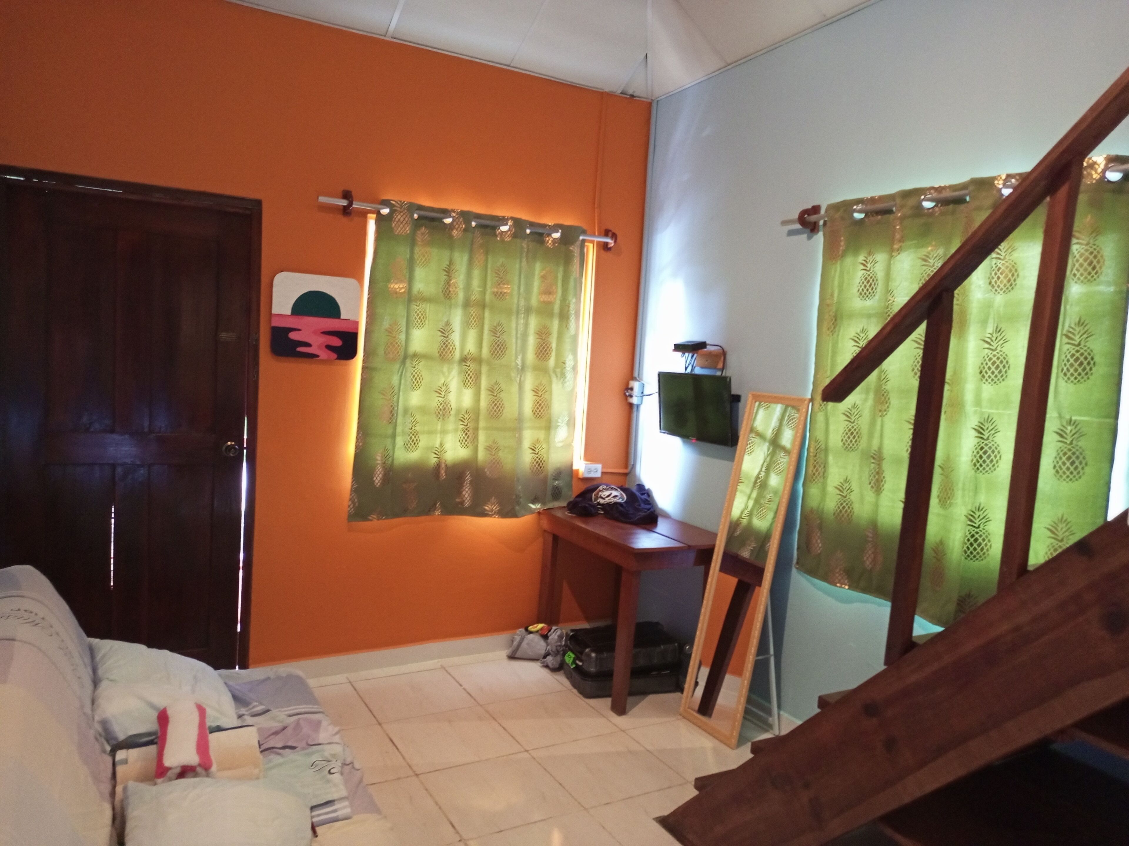 A room with an orange wall, a wooden door, a bed with pillows, a small table with a TV, and a staircase with a wooden railing.