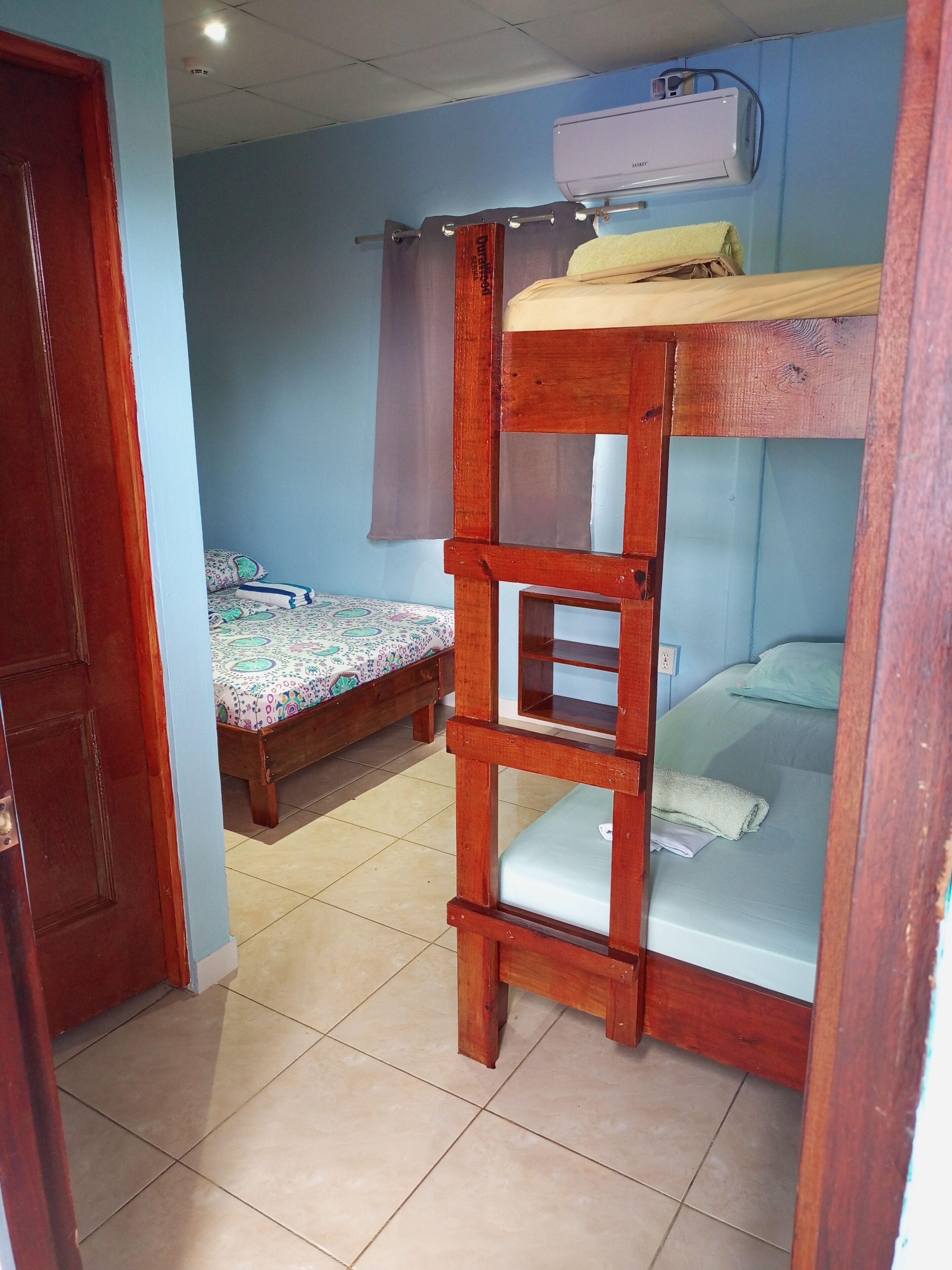 Une chambre avec un lit superposé, une échelle en bois, un lit recouvert d’une courtepointe à motifs et un appareil de climatisation fixé au mur.
