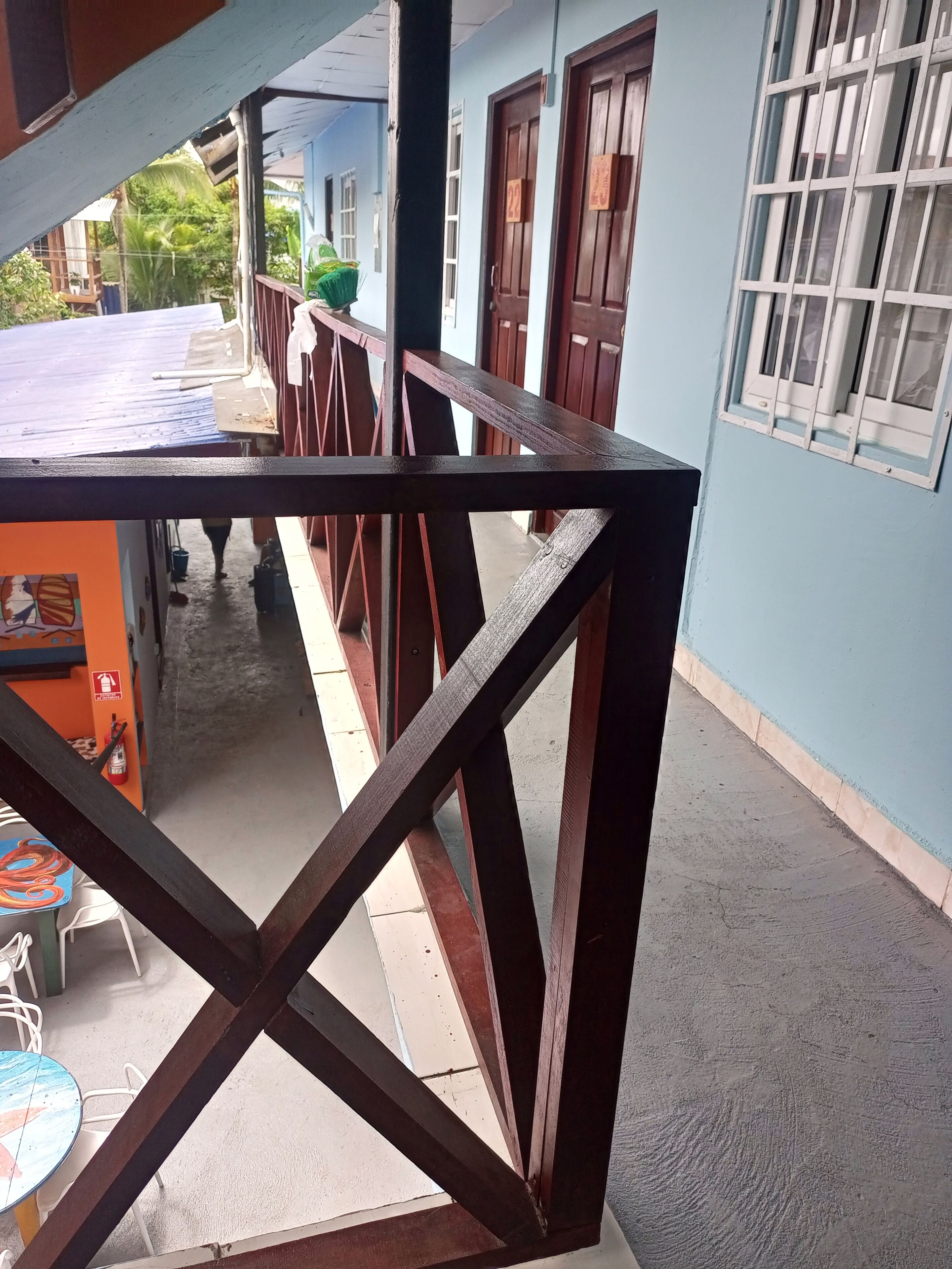 Un balcón con baranda de madera, una mesa con sillas y un edificio de paredes azules y puertas numeradas.