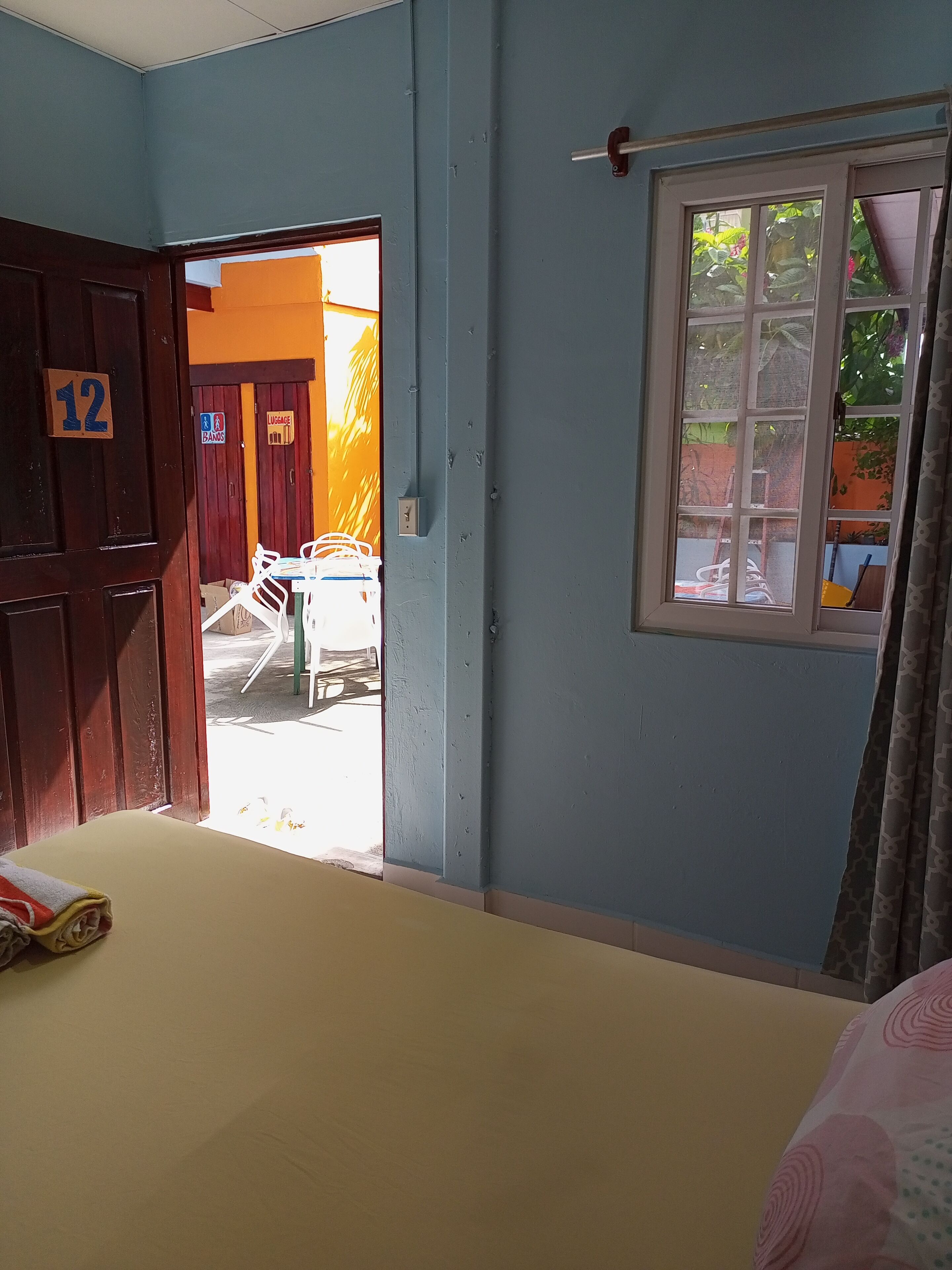 A room with a bed, a window with curtains, a door with a number, and a view of the outside.