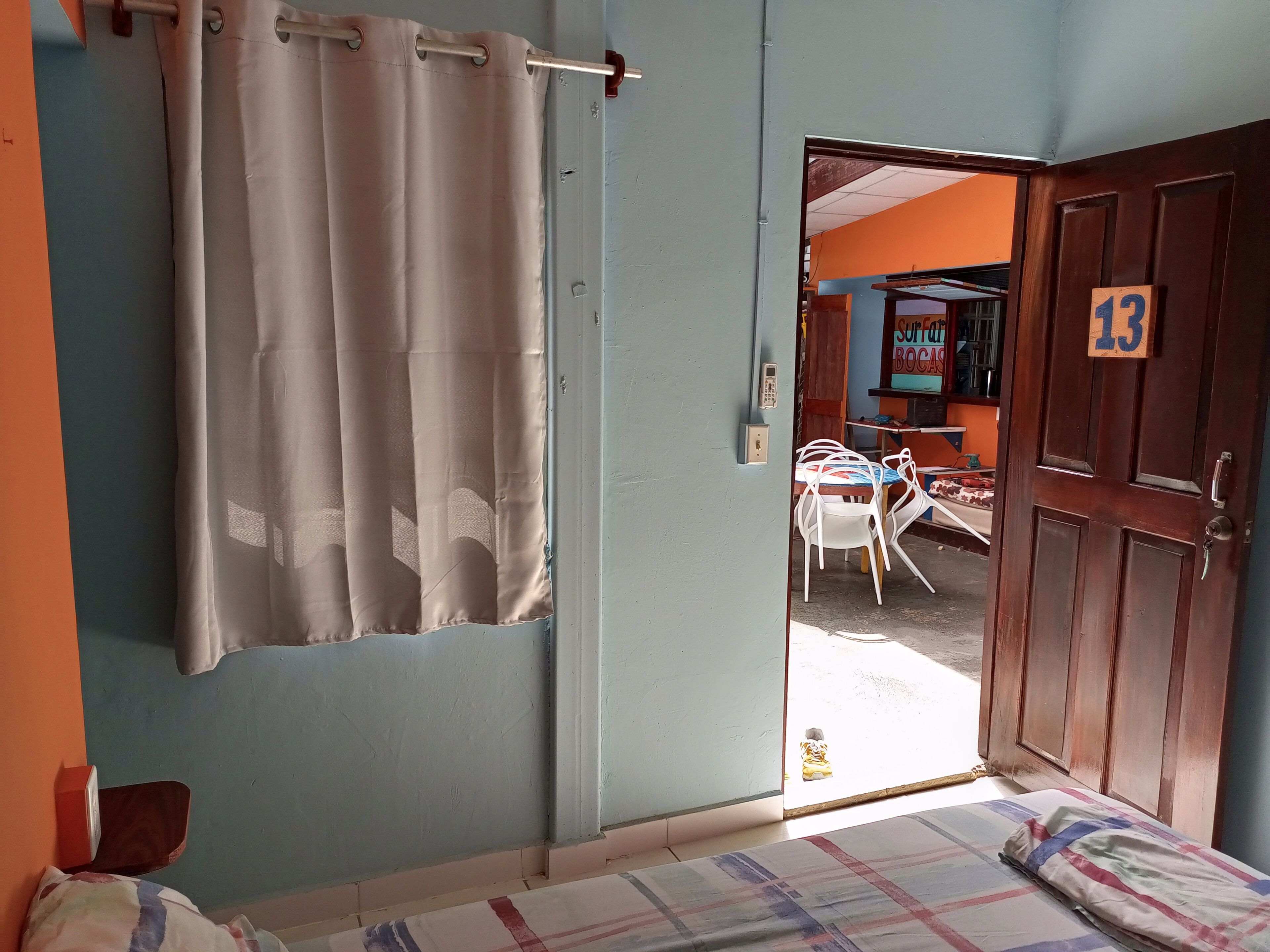 Une chambre avec un lit, un rideau, une porte en bois portant un numéro, et une vue sur la rue.