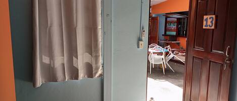 Une chambre avec un lit, un rideau, une porte en bois portant un numéro, et une vue sur la rue.