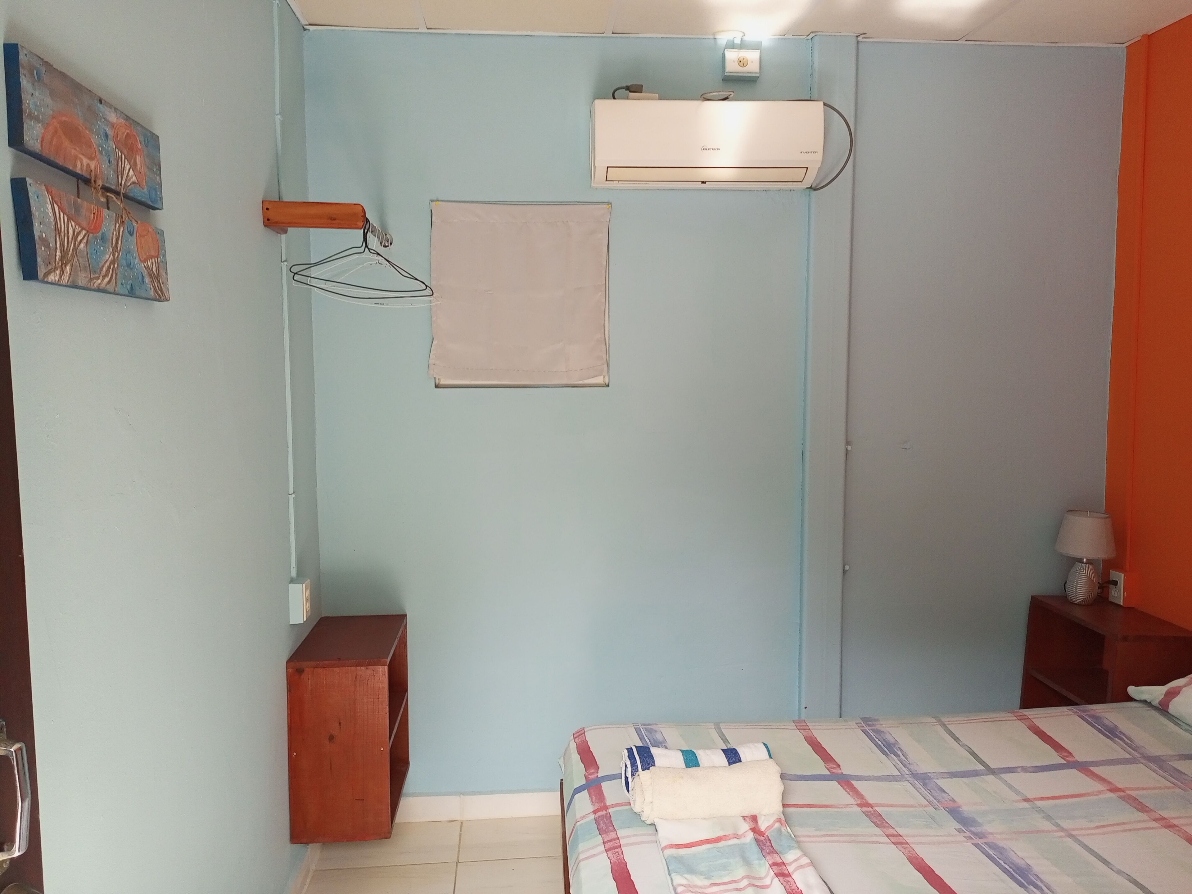 A bedroom with a bed, a wooden nightstand, a wall-mounted air conditioner, and a small shelf.