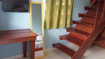 A room with a wooden staircase, a TV mounted on the wall, a wooden table, and a bed with a mirror.