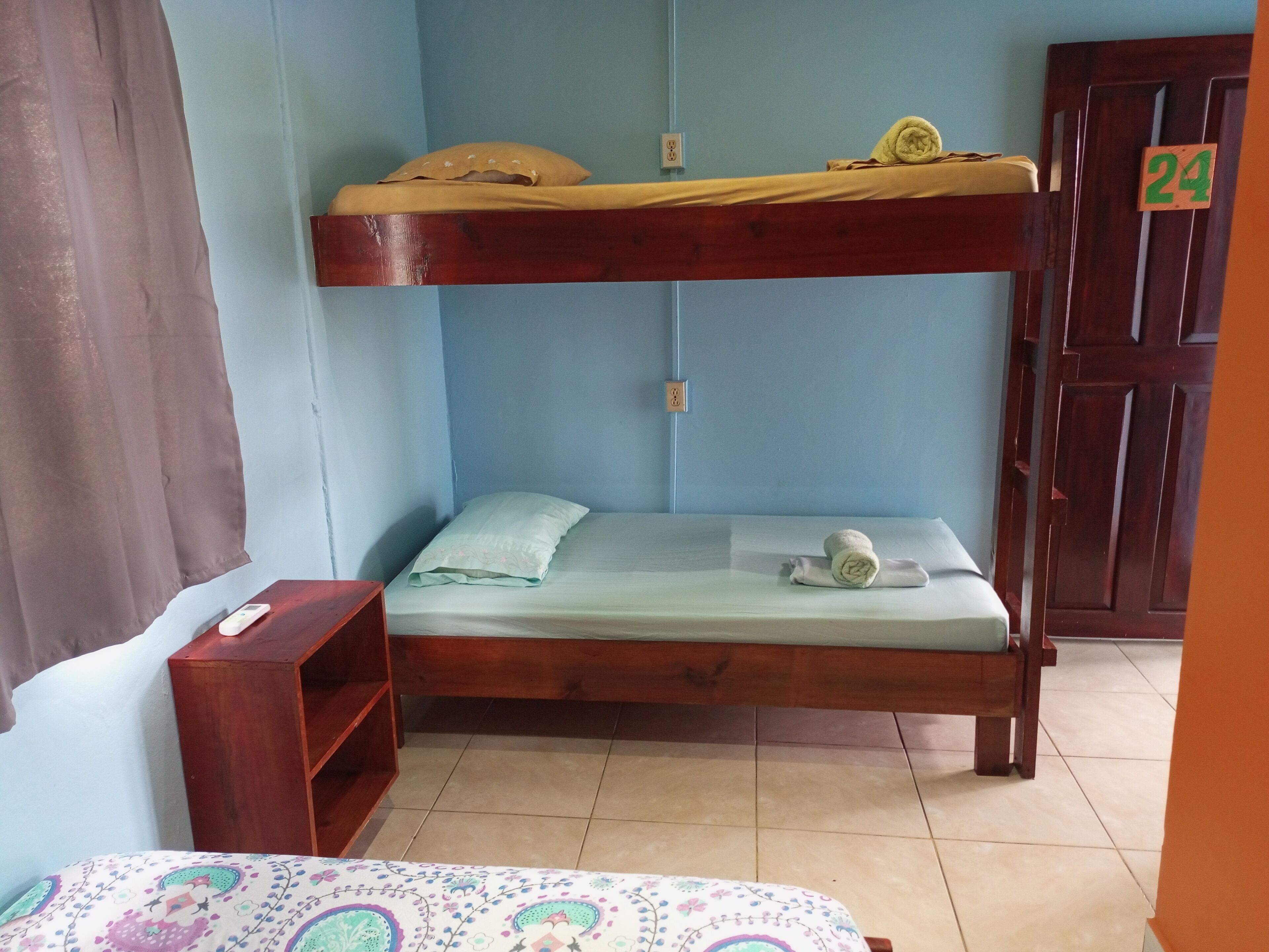 A bunk bed with a wooden frame, a lower bed with a light-colored mattress and a pillow, a small wooden shelf, and a door with a number sign.