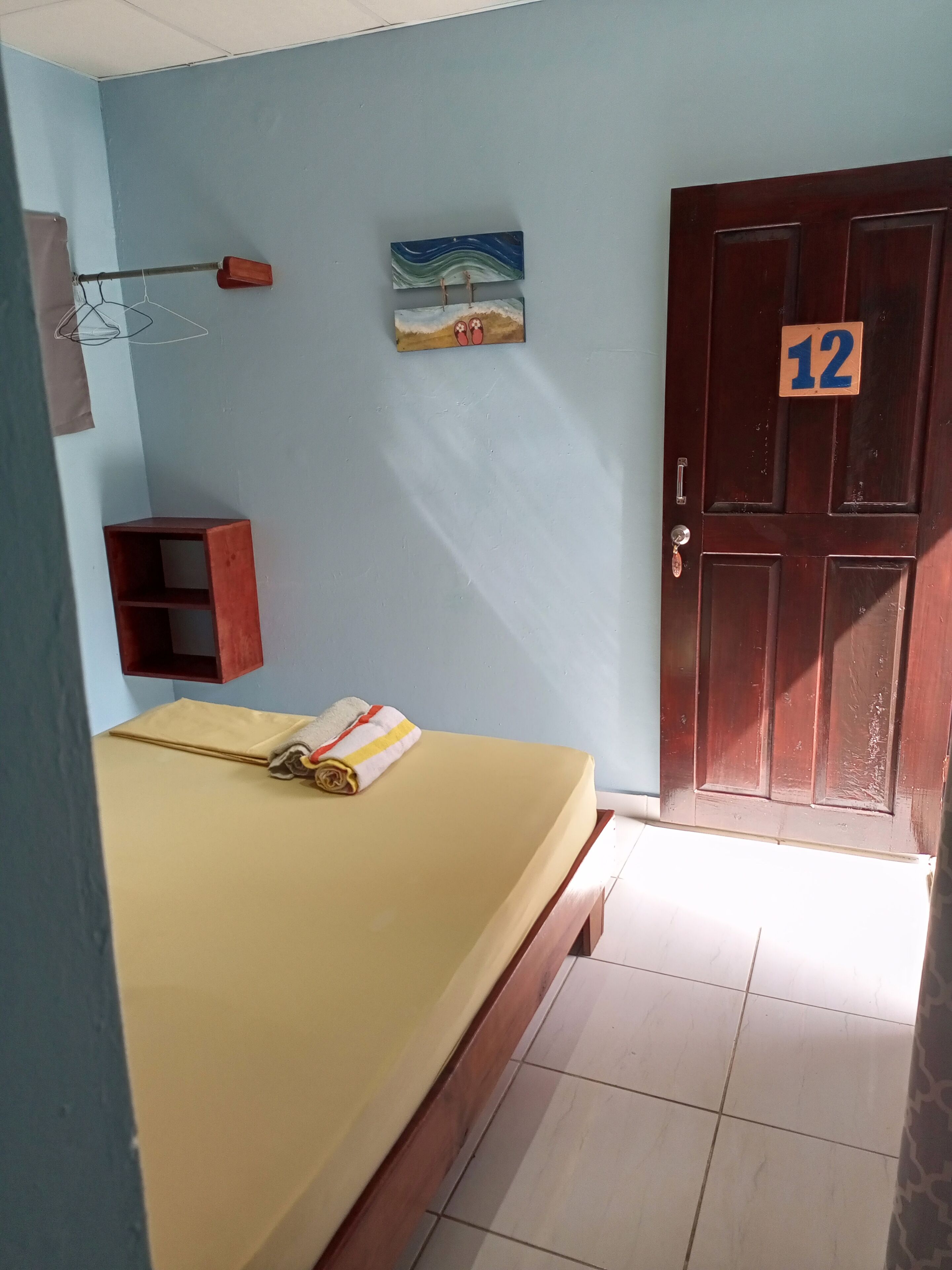 Una habitación con una cama, una puerta de madera con el número 12, un estante pequeño y un cuadro en la pared.