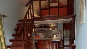 Un interior acogedor con una escalera de madera que conduce a un nivel superior, una zona de cocina con barra y un espacio de estar con sofá y cojines.