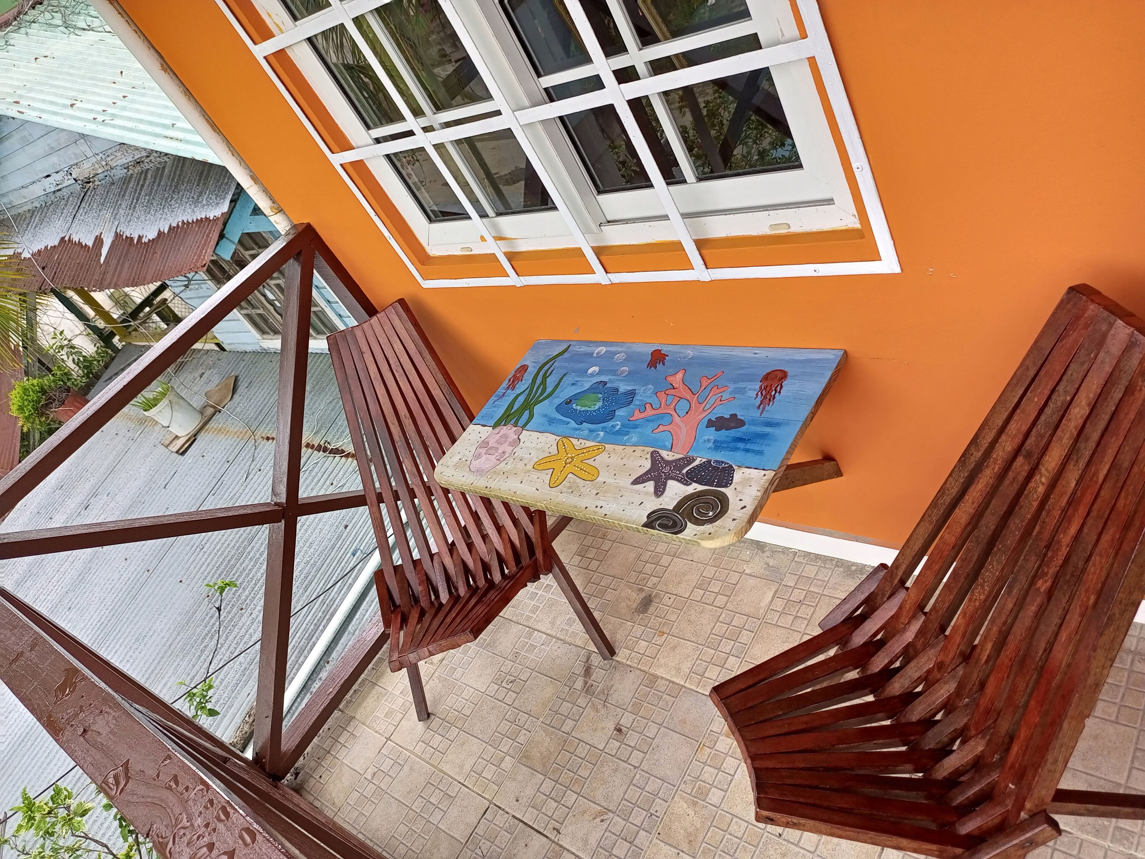 A balcony with wooden chairs, a small table with a painted artwork, and a window with white frames.
