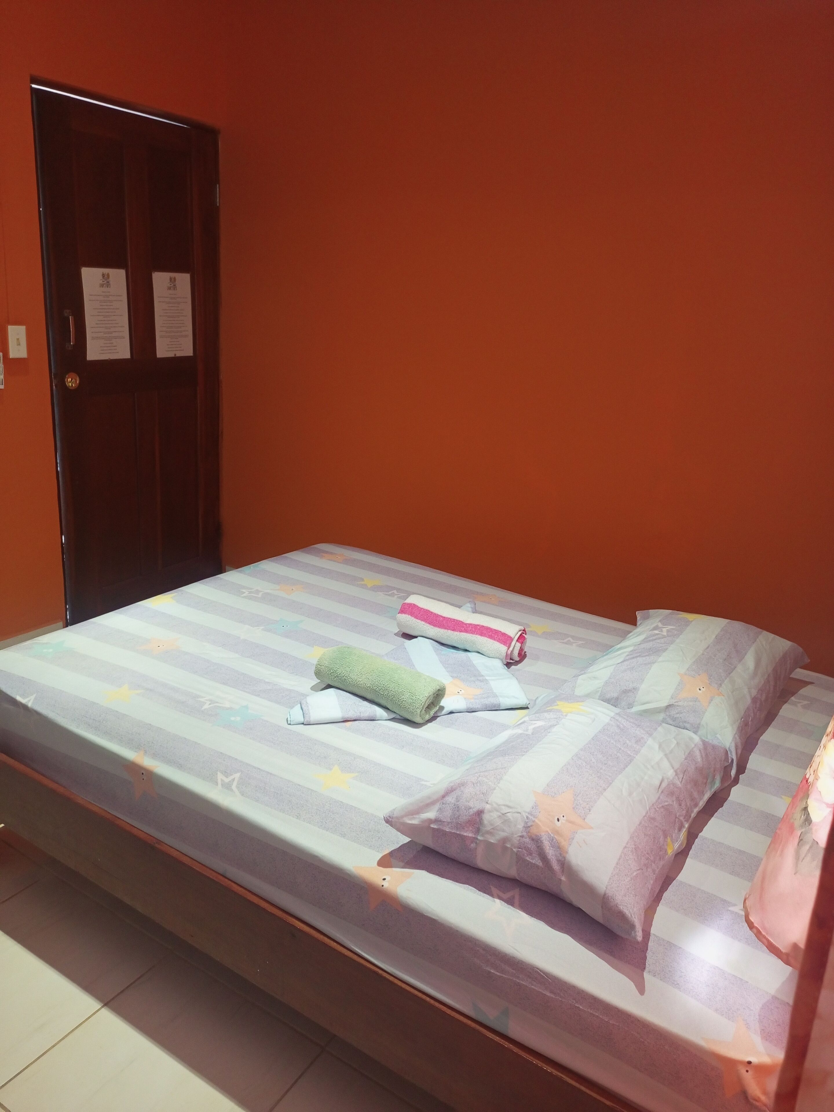 A single bed with a striped bedspread and pillows, a wooden door with a notice, and a tiled floor.