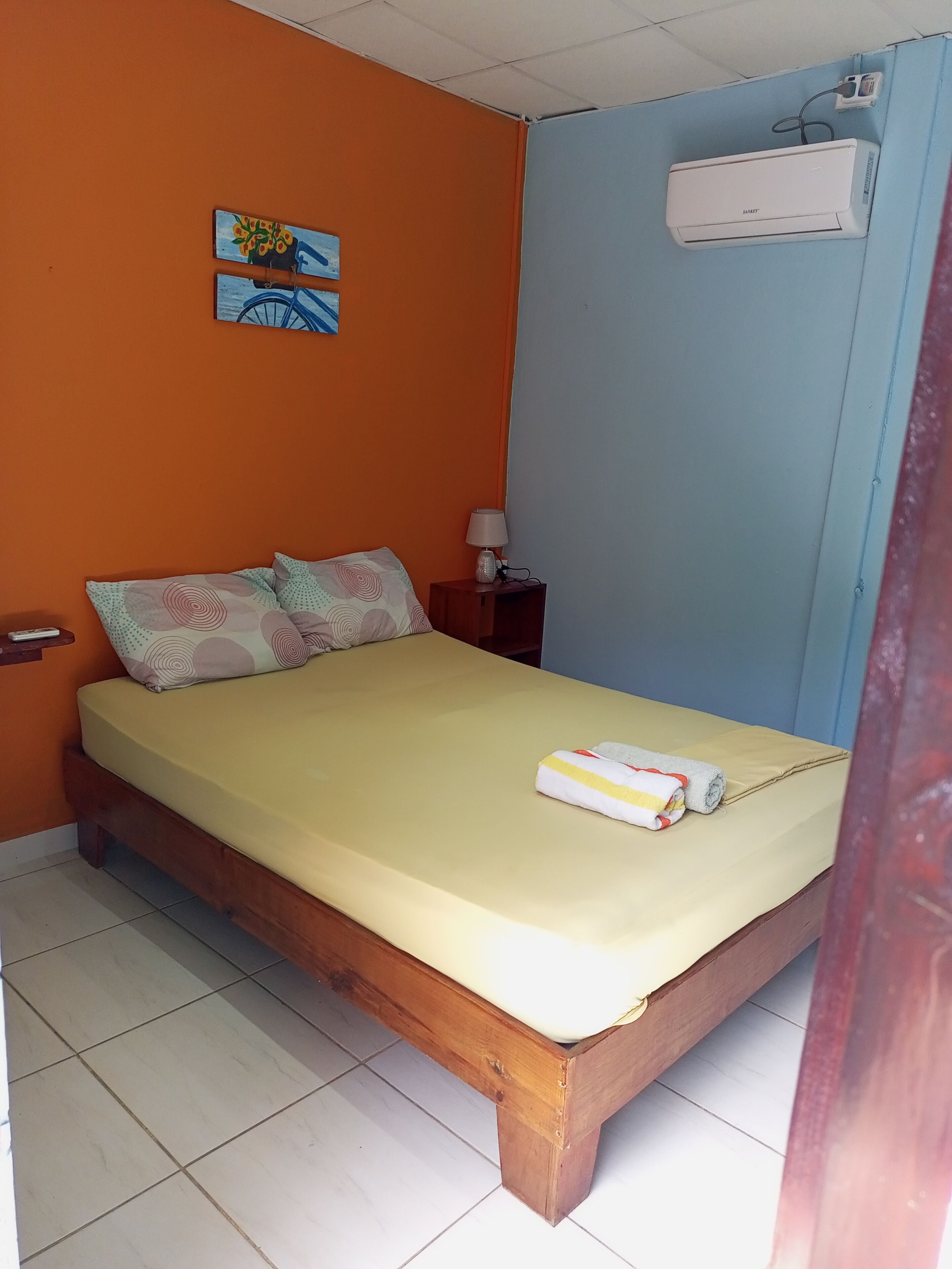 Una cama individual con colchón amarillo, estructura de madera y almohada con diseño circular. Hay una mesita de noche pequeña con una lámpara y un enchufe. Un equipo de aire acondicionado está montado en la pared sobre la cama.