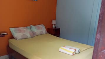 Una cama individual con colchón amarillo, estructura de madera y almohada con diseño circular. Hay una mesita de noche pequeña con una lámpara y un enchufe. Un equipo de aire acondicionado está montado en la pared sobre la cama.