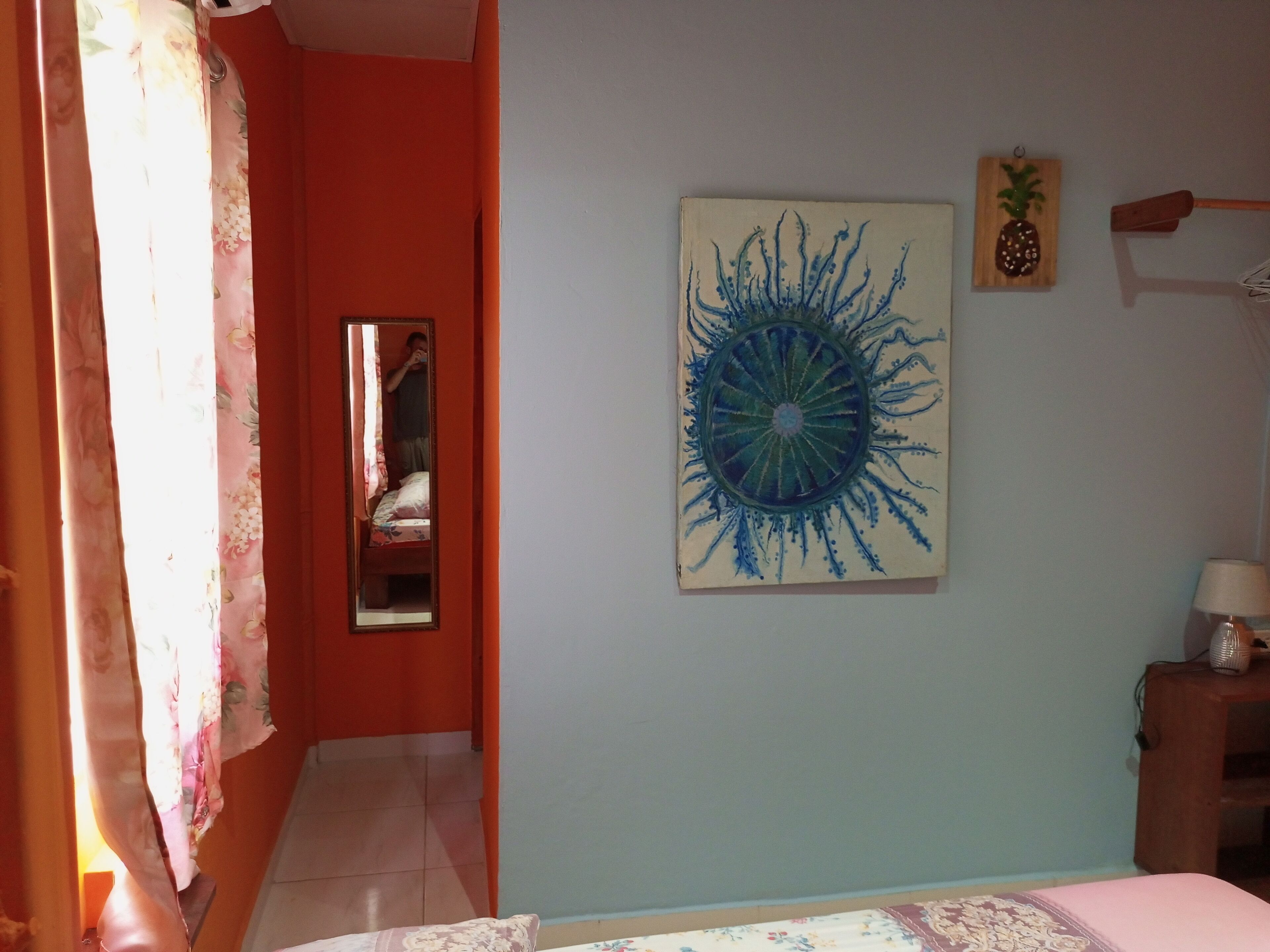 A bedroom with a bed, a mirror, a wall painting, and a small wooden shelf.