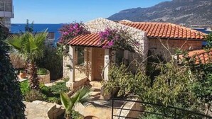 Un edificio de piedra con techo de tejas rojas, rodeado de vegetación y vistas al mar.