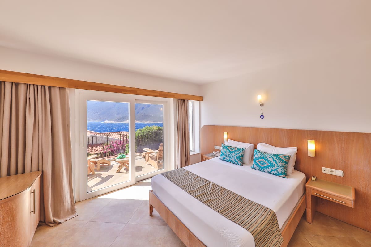 Una camera d'hotel con un grande letto, una testiera in legno, un comodino e vista sull'oceano attraverso una porta scorrevole in vetro.