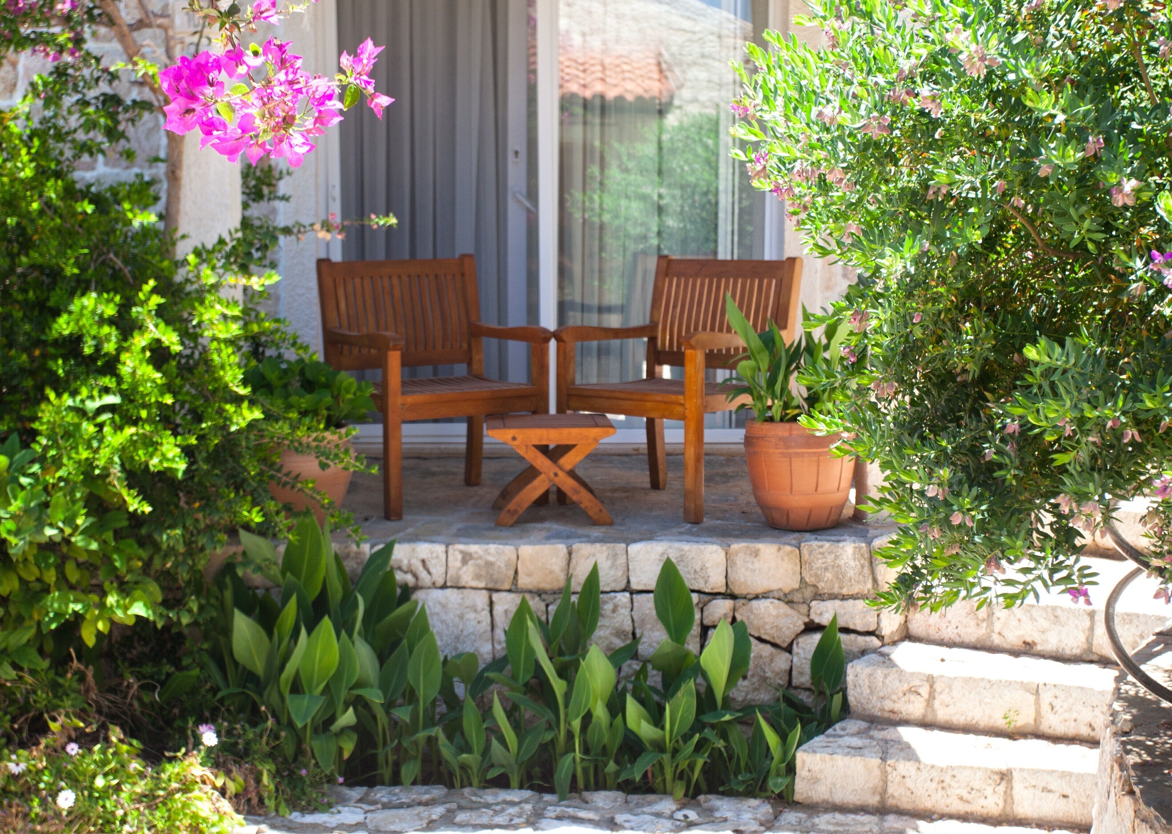 A wooden outdoor seating area with two chairs and a table, surrounded by potted plants and a stone wall.