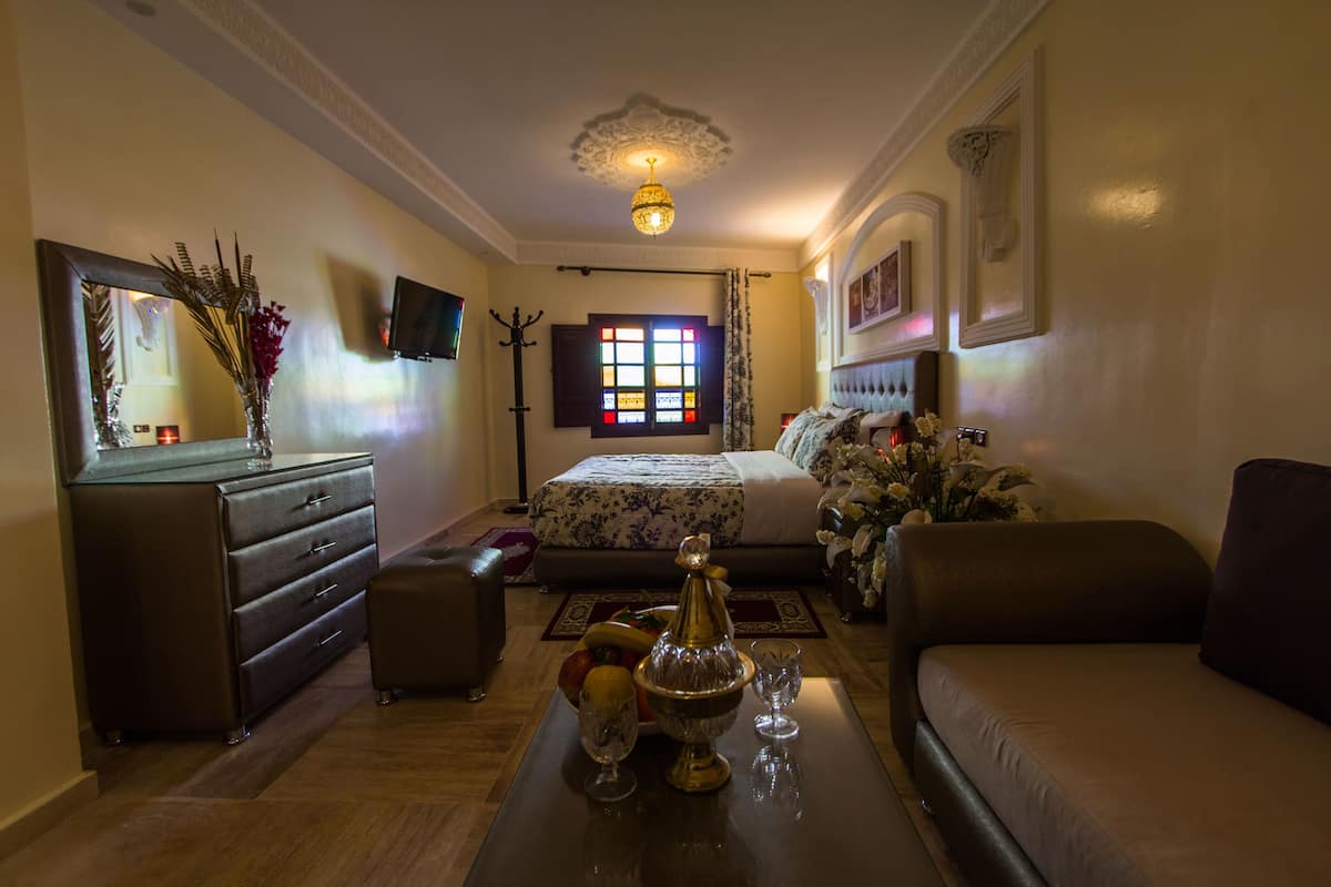 A hotel room with a bed, a sofa, a dresser, a mirror, a chandelier, and a window with curtains.