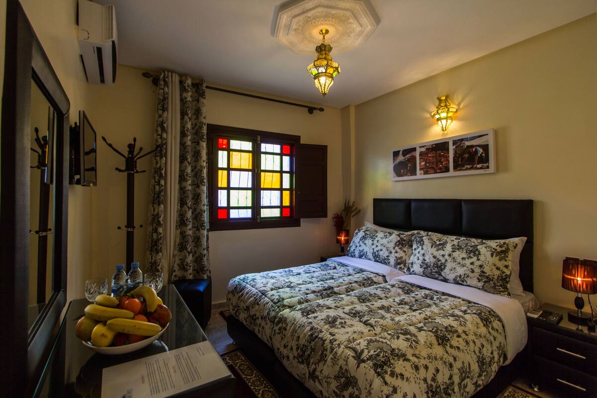A hotel room with a bed, a bedside table with a lamp, a window with curtains, and a wall-mounted picture.