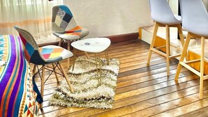 A cozy room with a wooden floor, a small round table, two chairs, a colorful sofa, and a large window with sheer curtains. - Blue Door Housing Historic Quito (Quito)