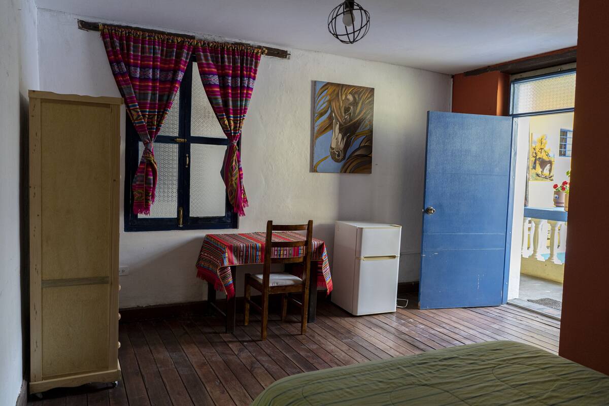 Ein Zimmer mit einem Holzschrank, einem Tisch mit bunten Tischdecken, einem Stuhl, einem kleinen, weißen Kühlschrank und einer blauen Tür.