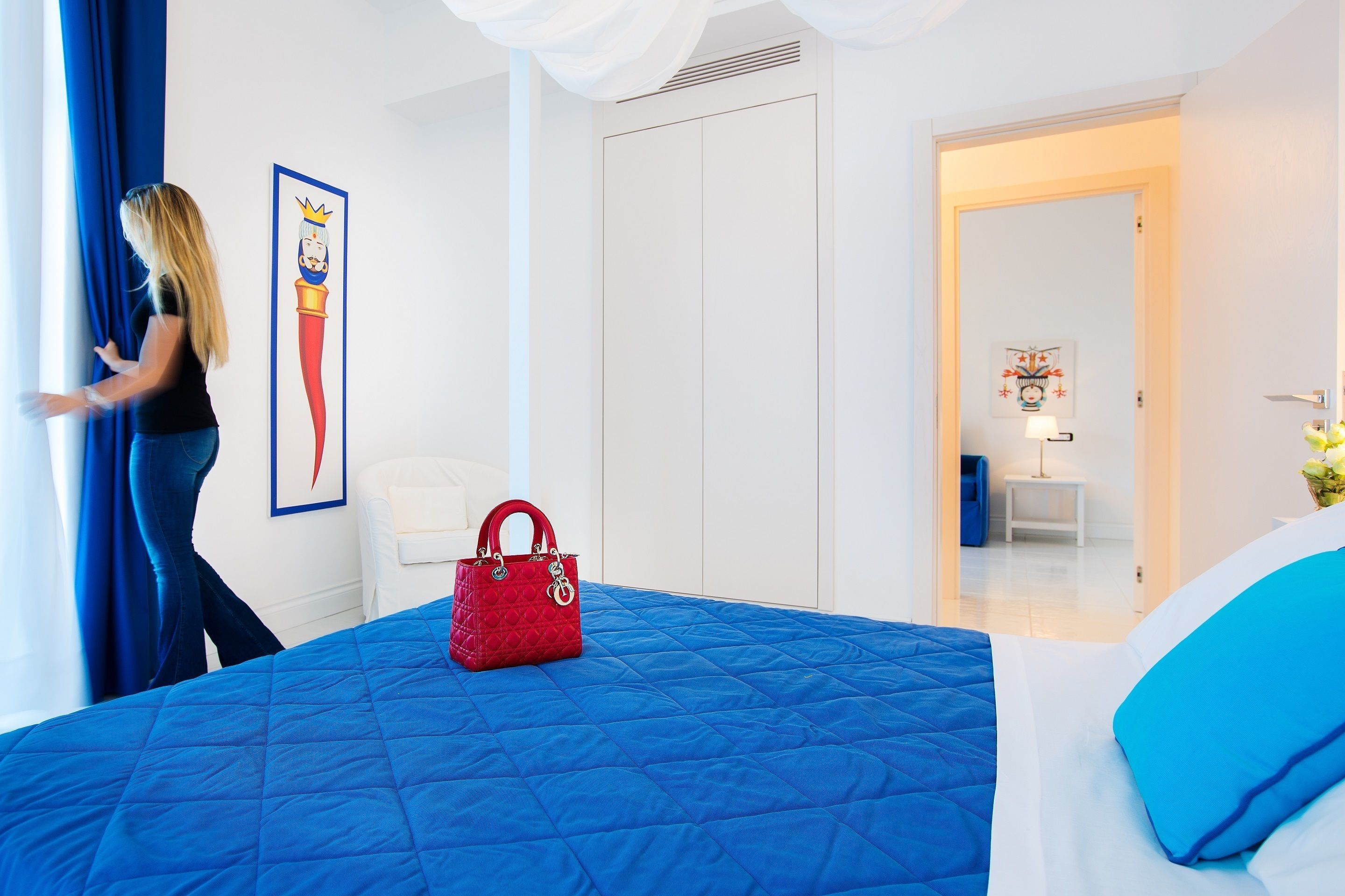 Une chambre d’hôtel avec un lit, un sac à main rouge, une chaise blanche et un rideau bleu.