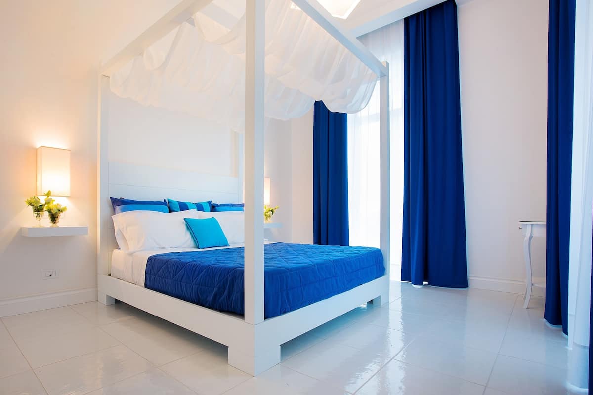 A four-poster bed with a white frame and blue bedding, a bedside table with a lamp, and a view of the room's white walls and floor.