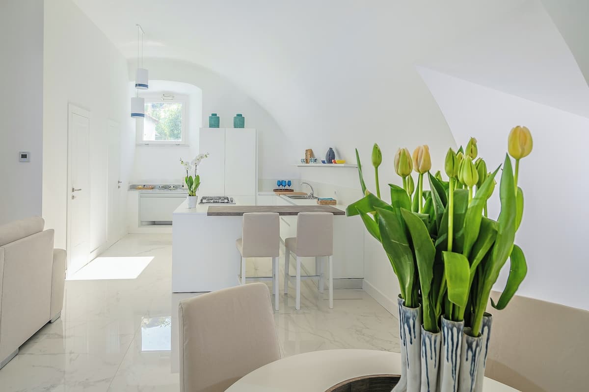 A modern kitchen with white cabinets, a central island, and a dining area with two chairs. There is a vase with green plants in the foreground.