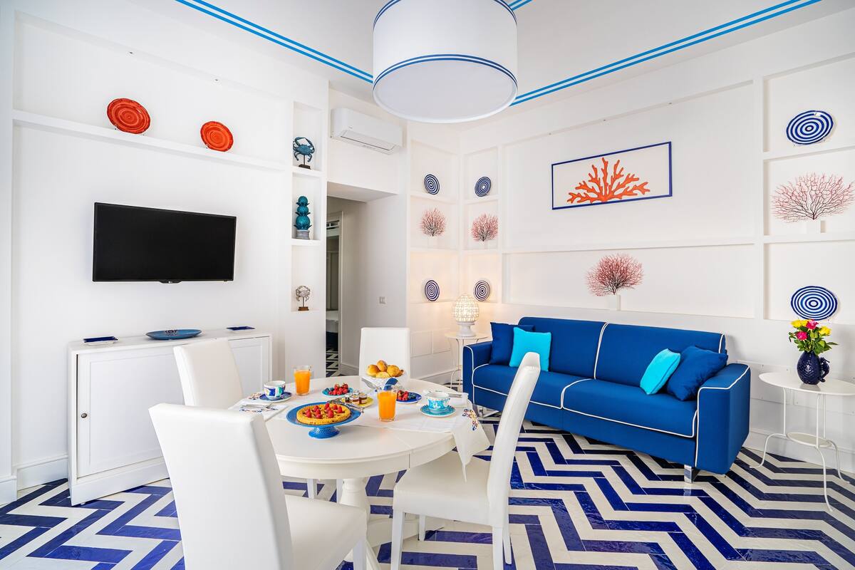 A modern living room with a blue sofa, white dining table, and a flat-screen TV.