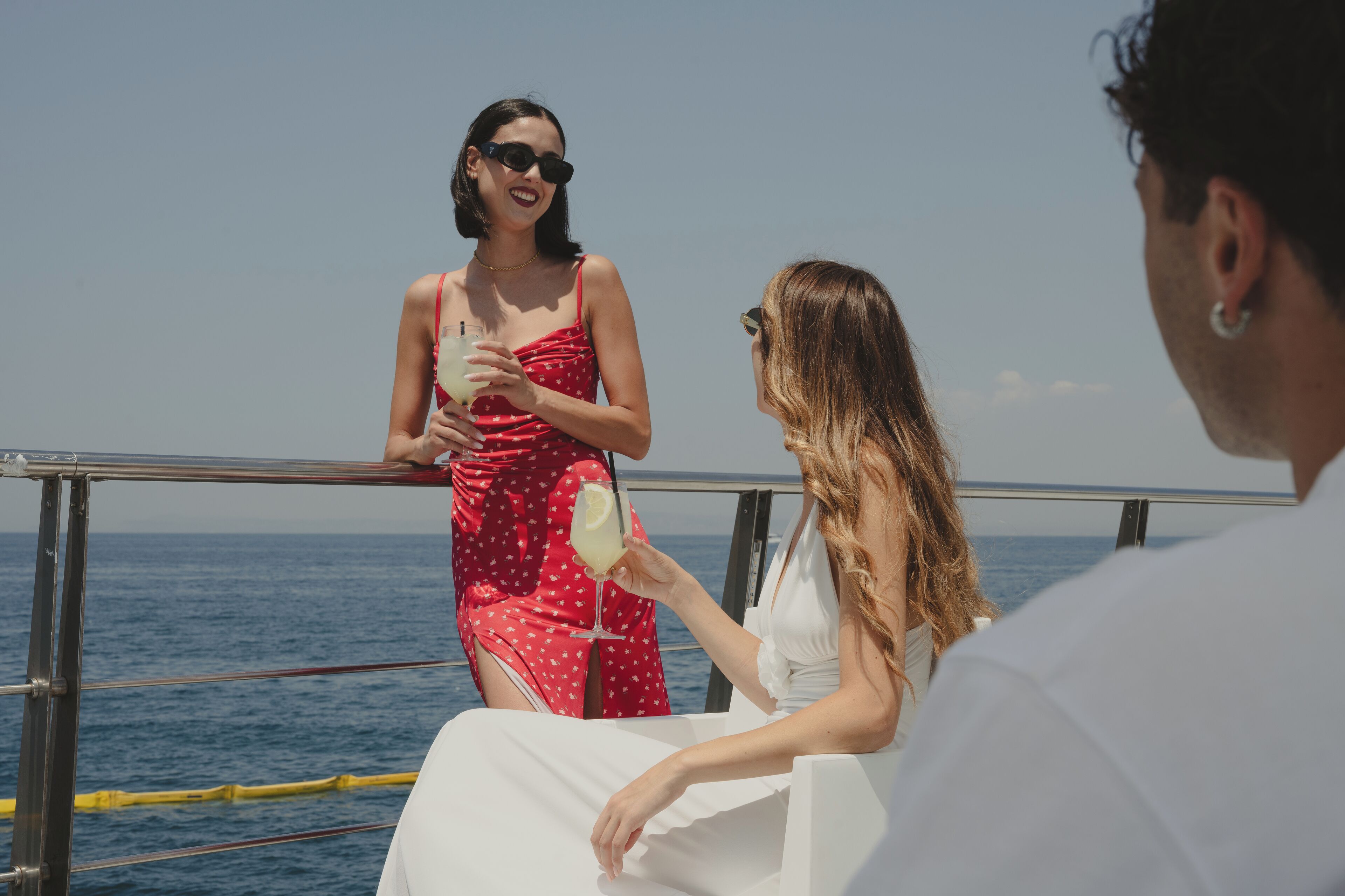 A person in a red polka dot dress holding a drink on a boat with a railing and ocean view.