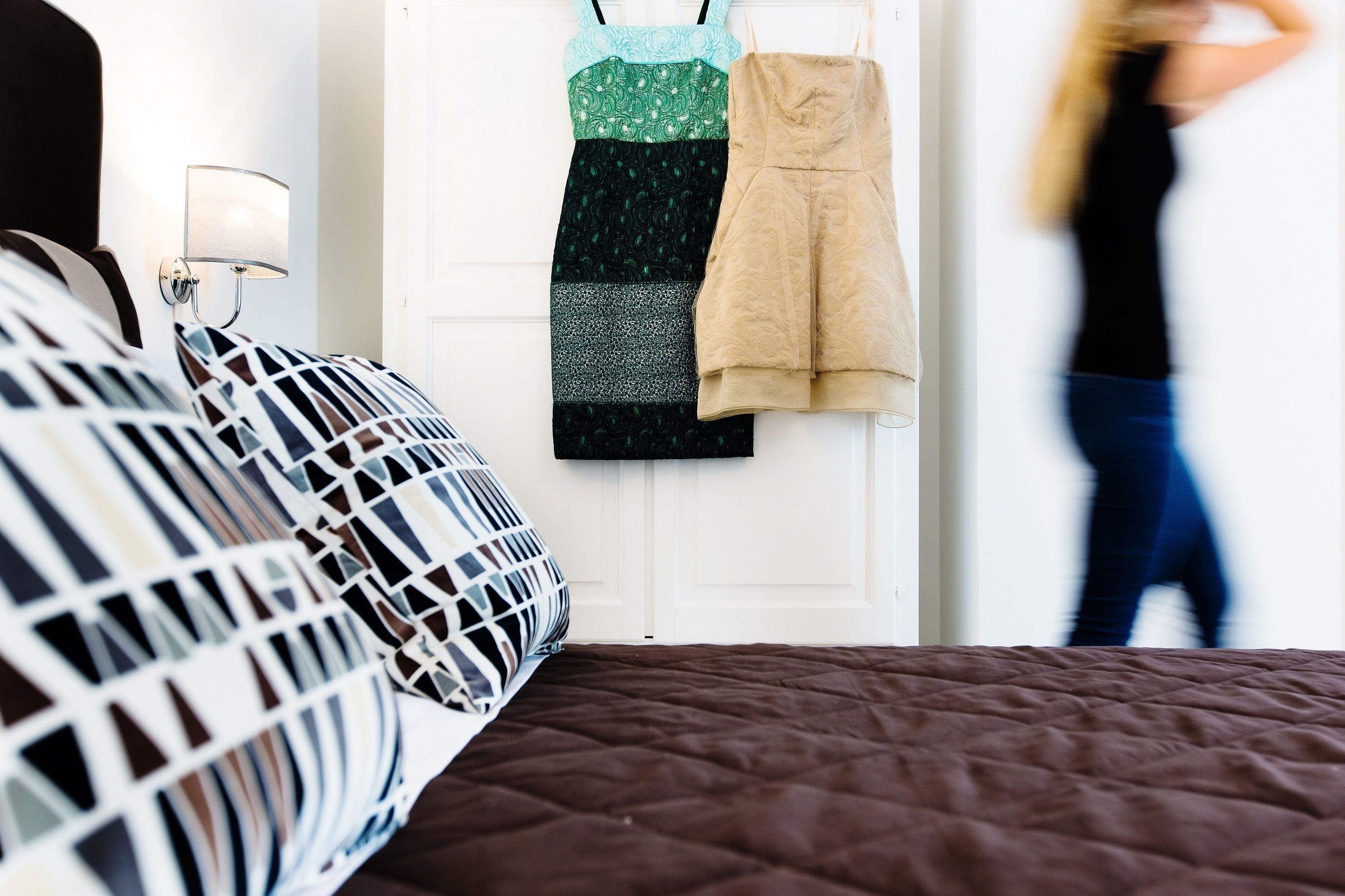 Un lit avec un oreiller à motifs et une courtepointe, une lampe fixée au mur, et une porte sur laquelle sont accrochées deux robes.