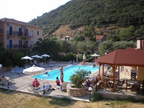 Zona de piscina amb gandules i ombrel·les, envoltada d'edificis i vegetació.
