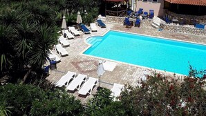 A swimming pool with lounge chairs and umbrellas, surrounded by greenery and buildings. - Hotel Nostos (Ithaki)