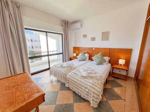 A hotel room with a large bed, two bedside tables, a wooden bench, a balcony with curtains, and an air conditioning unit. - Cerro Atlantico by Portugalferias (Albufeira)
