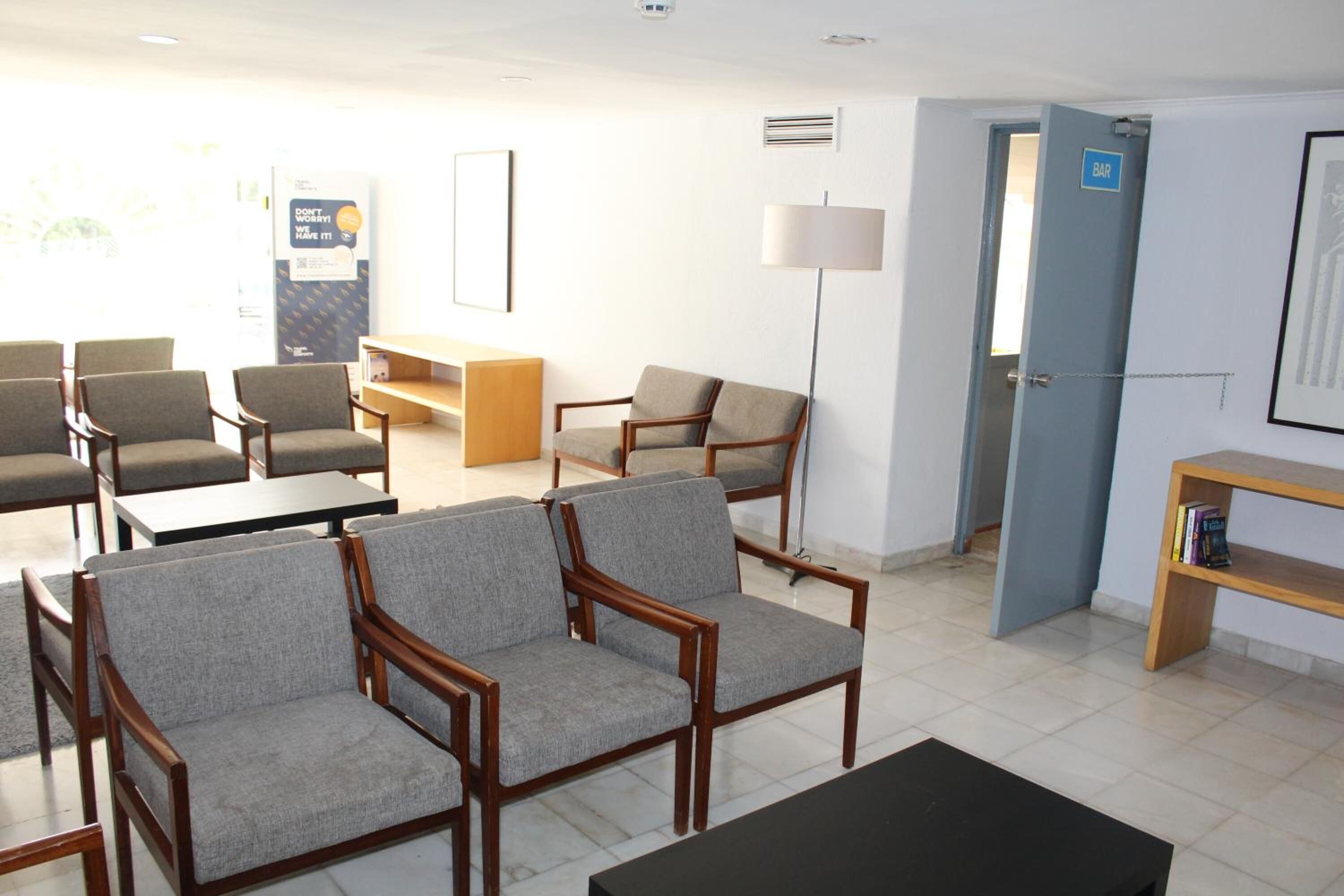 A waiting area with grey upholstered chairs, a wooden table, a lamp, and a bookshelf.
